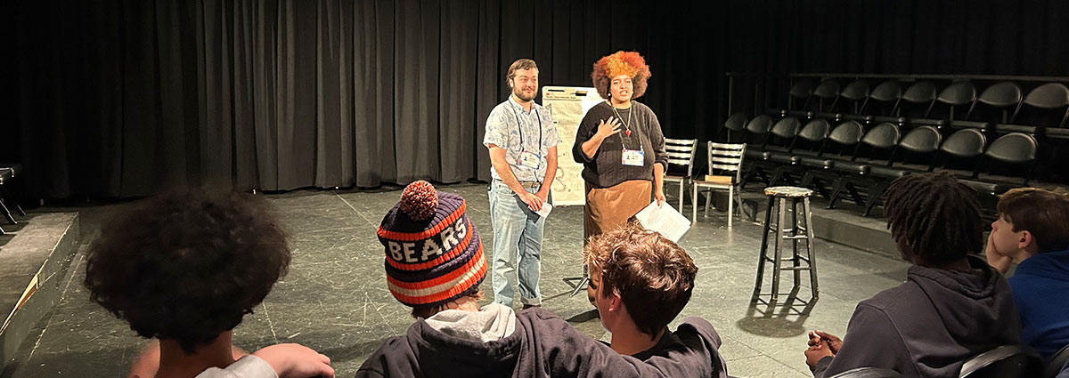 Two workshop facilitators stand onstage in a black box theatre talking to an audience